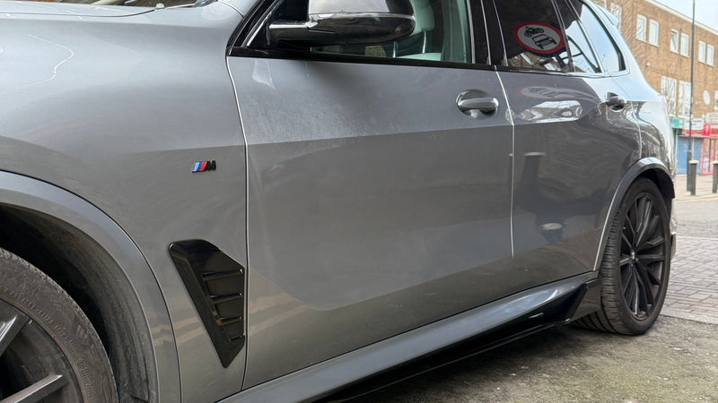 A gray SUV is parked under a concrete structure beside a building. It features a black M badge on the side, indicating its model variant.