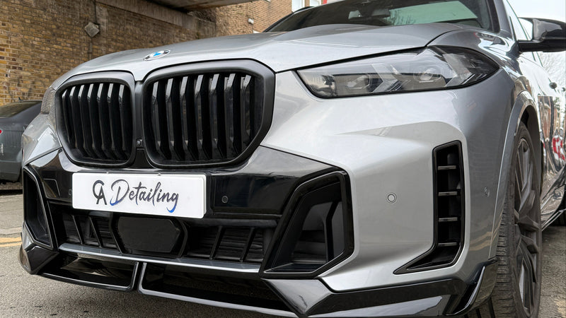 A silver BMW with a prominent grille is parked under an urban bridge. A sign reads "A Detailing." The setting includes brick walls and street signs.