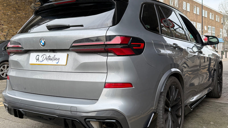 A silver BMW SUV is parked under a bridge, displaying "A Detailing" on the license plate. Nearby, brick buildings and street signs are visible in an urban setting.
