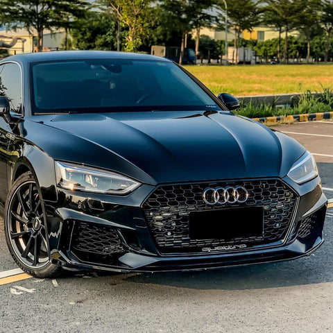 A sleek black Audi car is parked on a road, with a clear sky and green trees in the background. The front license plate reads "quattro."
