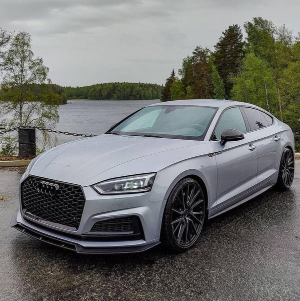 A silver sedan is parked on a wet road, surrounded by lush green trees and a lake in the background, creating a serene, natural setting.