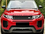 A red Range Rover is parked on a brick driveway, surrounded by lush greenery. The front of the car prominently displays the 