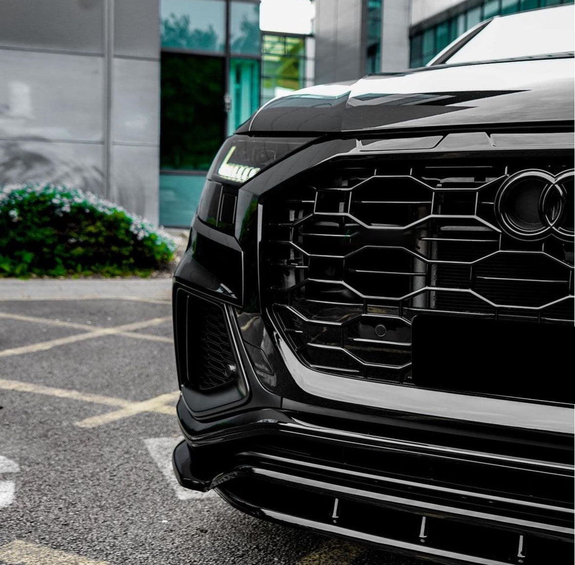 A black car's front with a hexagonal grille and LED headlights, parked in an urban parking area near modern glass buildings and greenery.