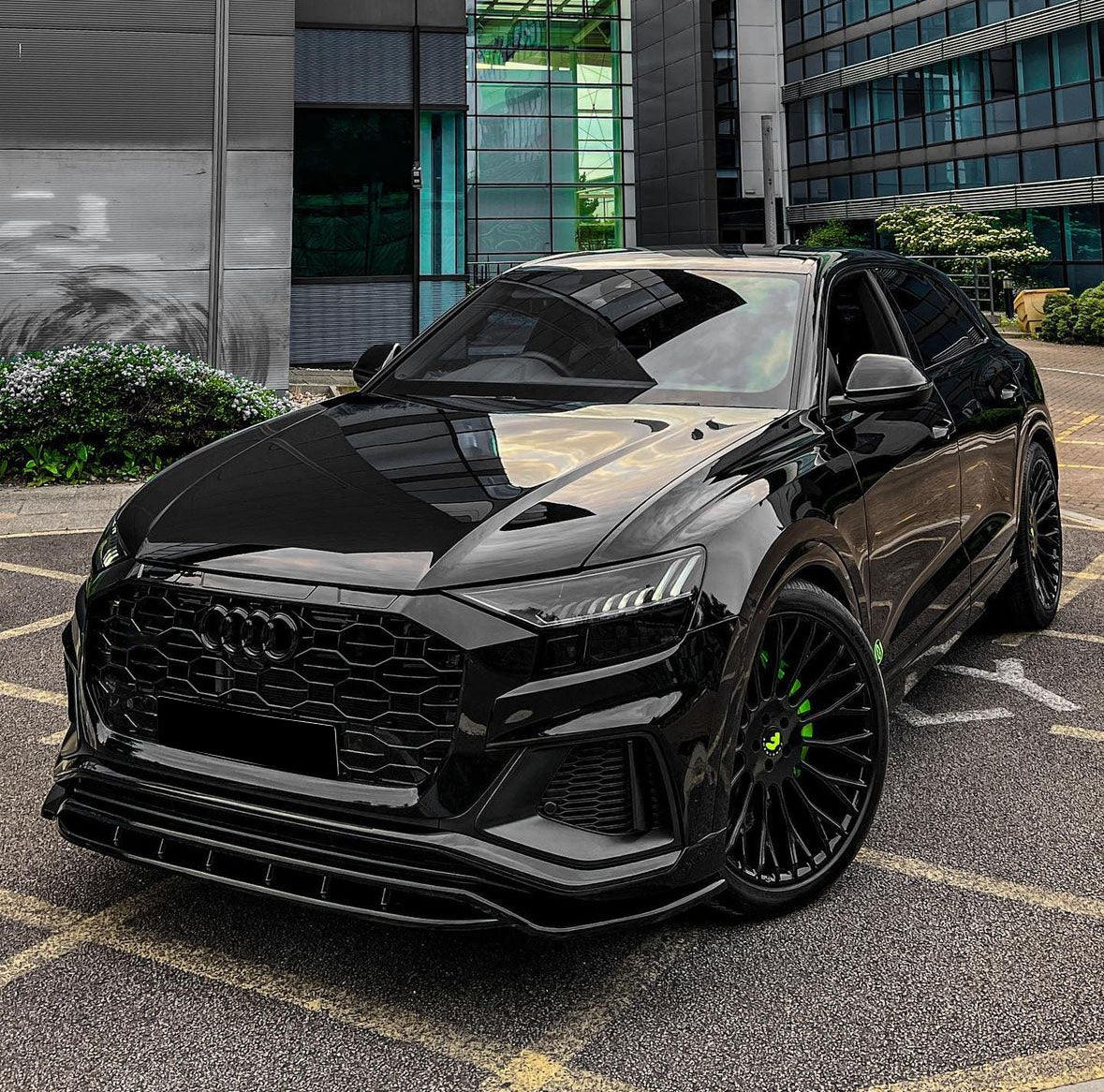 A sleek black car is parked on a lot, featuring green-accented wheels, in front of a modern glass and concrete building.
