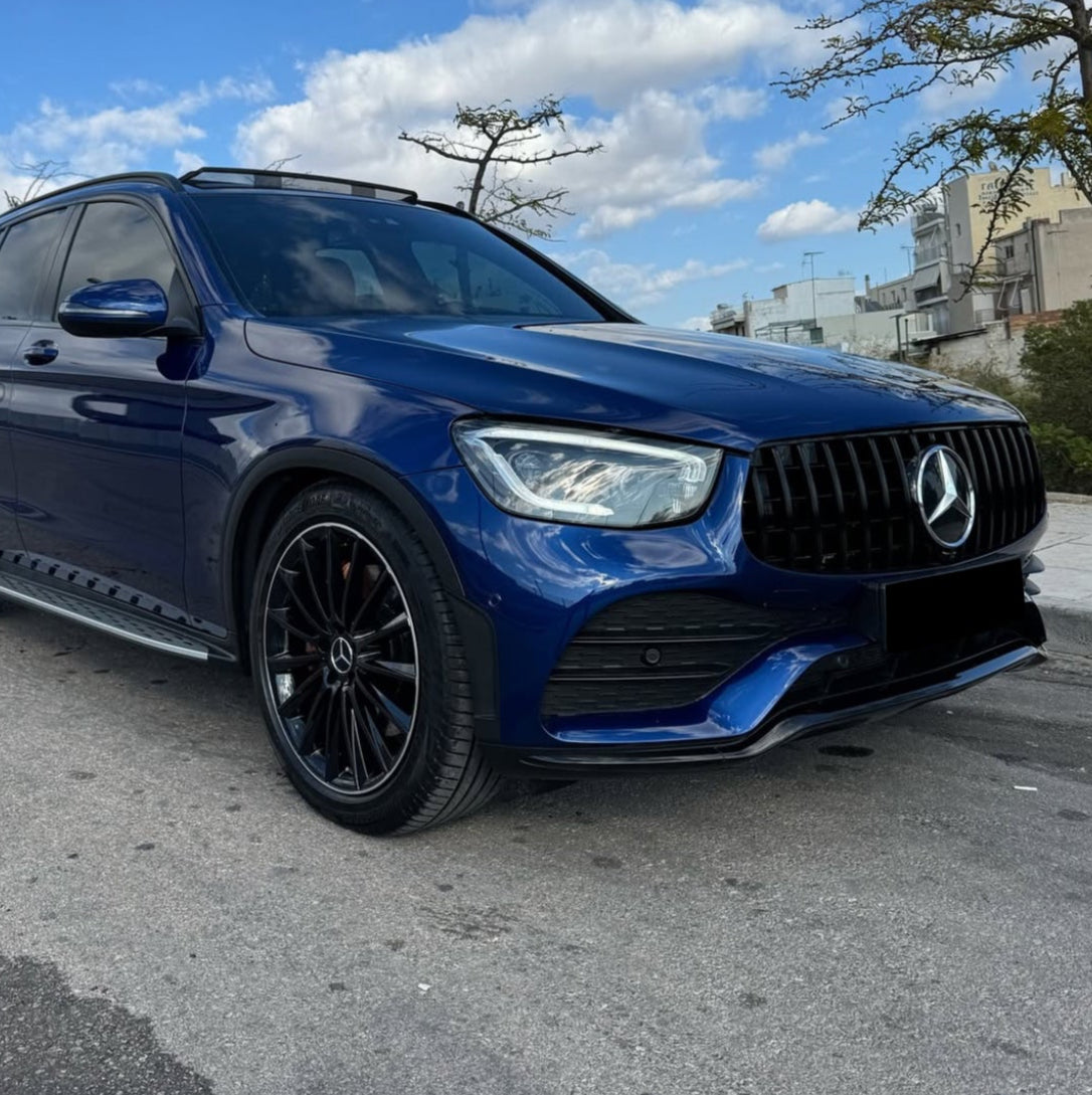 A blue Mercedes-Benz SUV is parked on a street, featuring black alloy wheels and a prominent front grille. The background includes a clear sky with scattered clouds and urban buildings.
