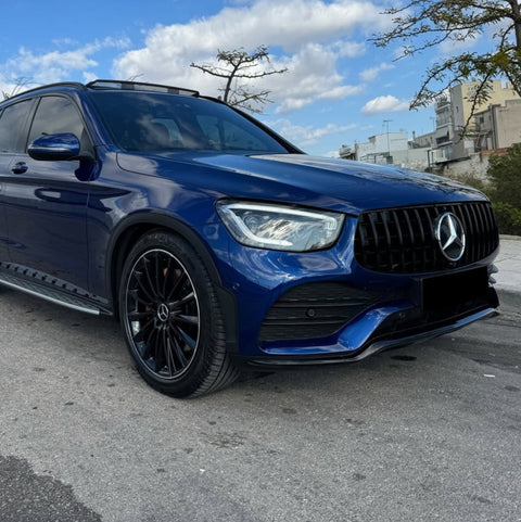 A blue Mercedes-Benz SUV is parked on a street, featuring black alloy wheels and a prominent front grille. The background includes a clear sky with scattered clouds and urban buildings.