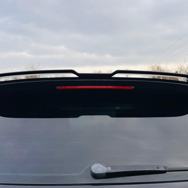 A car with a rear window featuring a wiper and a spoiler, surrounded by overcast skies and leafless trees.