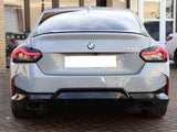 A gray BMW 220i is parked on a brick driveway. The car's rear view features sleek taillights and a visible badge. A building with glass doors and windows surrounds it.