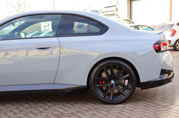 A gray coupe is parked on a brick surface, featuring black alloy wheels and red brake calipers. Other vehicles and a building are visible in the background.