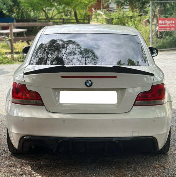 A white BMW car is parked, reflecting tree silhouettes on its rear window. It is situated on a gravel path near a "Wellgate Community Farm" sign among greenery and a wooden bench.