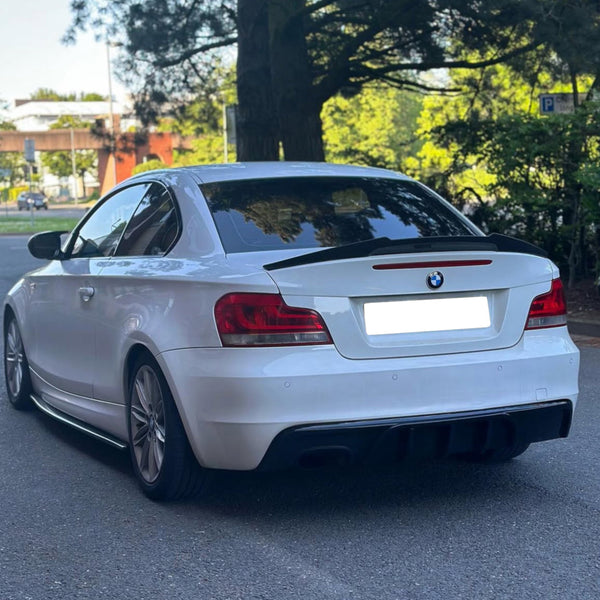 A white car is parked on a paved area, surrounded by trees and foliage, under a clear, sunny sky. The license plate is obscured.