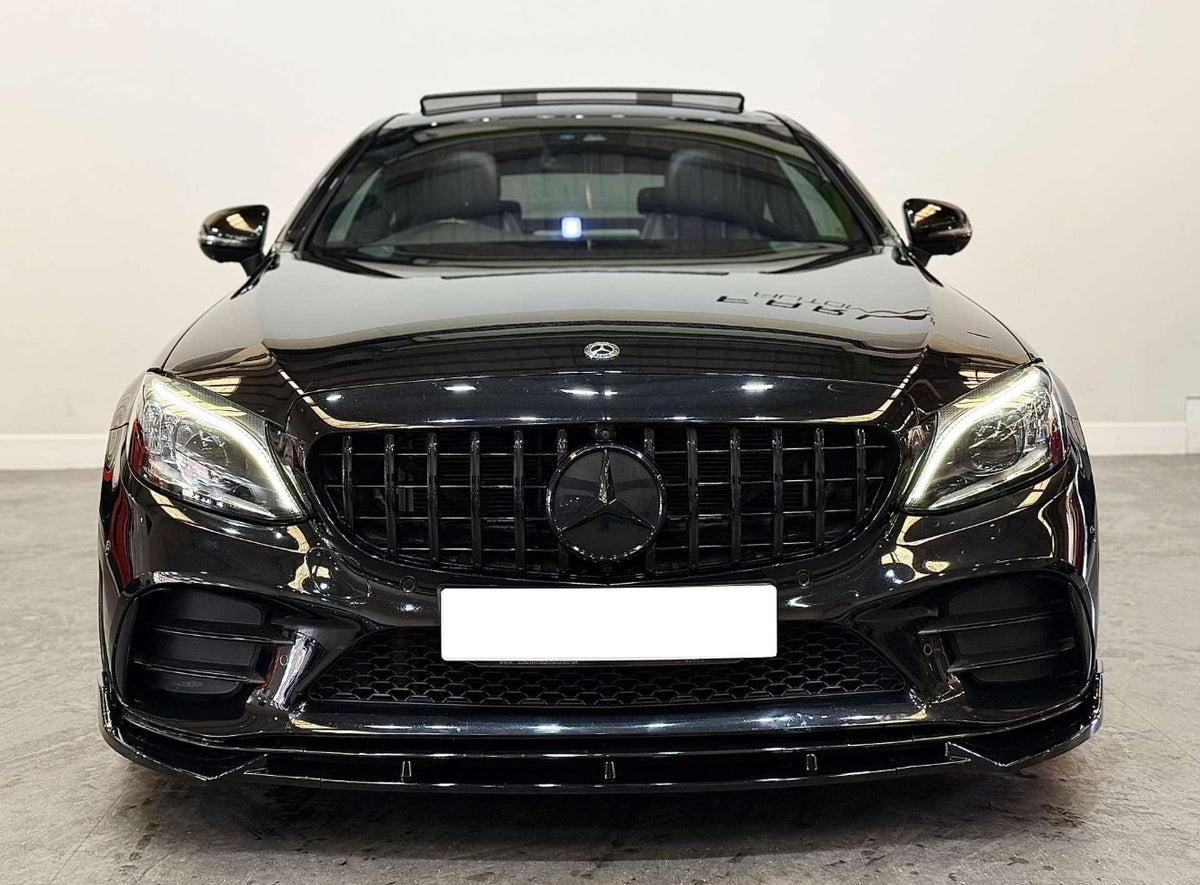 A black luxury sedan with illuminated headlights is parked on a concrete floor in a white-walled room. The license plate is blank, and a Mercedes-Benz emblem is visible.
