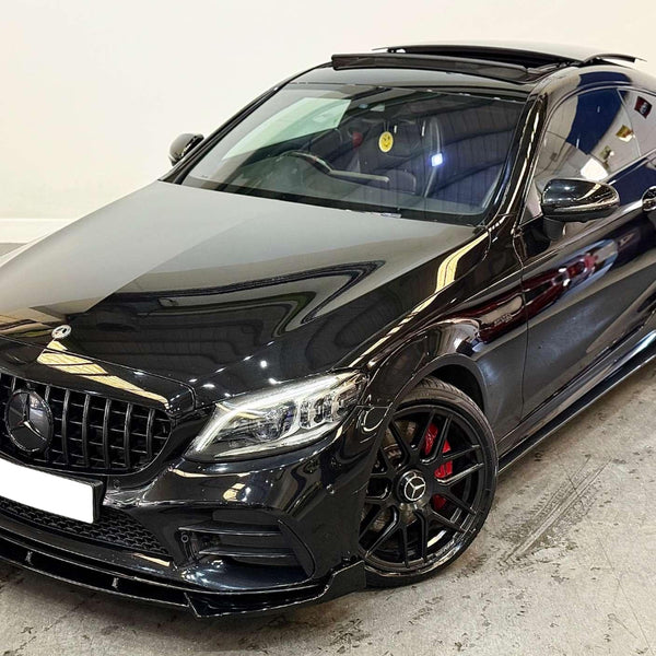 A black Mercedes-Benz coupe with an open sunroof is parked indoors. It features a sleek design, AMG wheels with red brake calipers, and a smiley face air freshener inside.