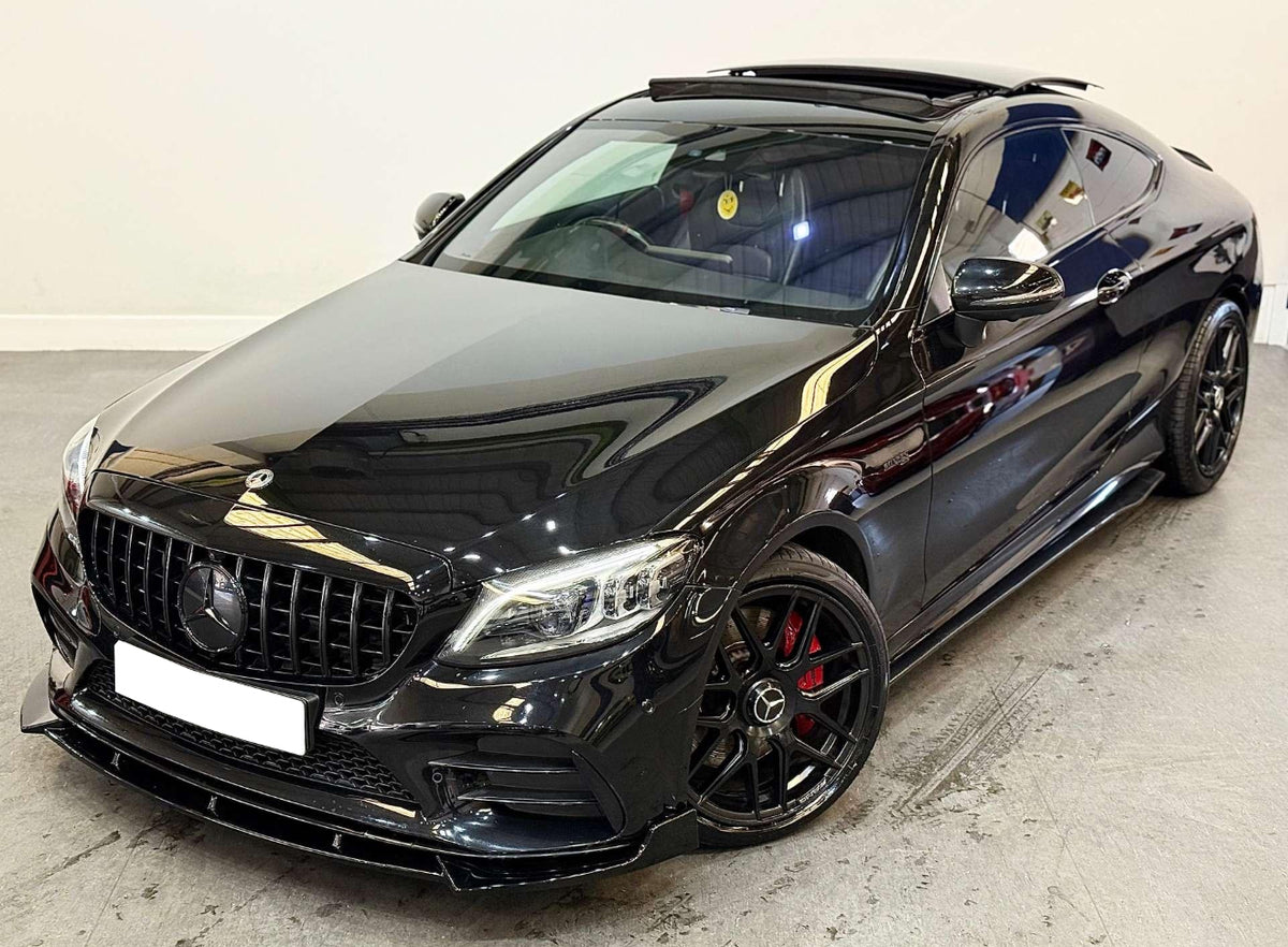 A black Mercedes-Benz coupe with an open sunroof is parked indoors. It features a sleek design, AMG wheels with red brake calipers, and a smiley face air freshener inside.