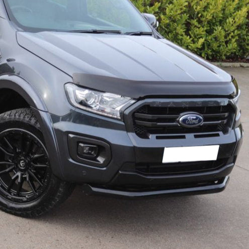 A dark gray truck with a "Ford" emblem on the front grille is parked on a concrete road, surrounded by green bushes. The headlights are sleek and modern.