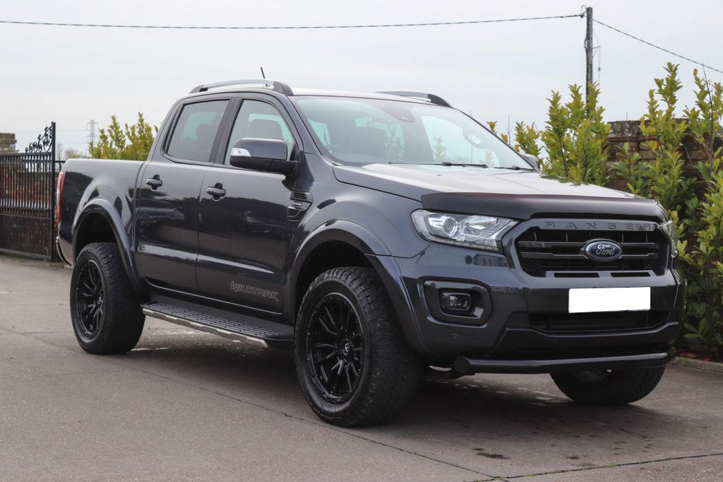 A black pickup truck is parked on a concrete surface, surrounded by greenery and a metal fence. The vehicle has "Wildtrak" branding on its side.