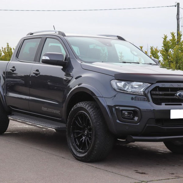 A black pickup truck is parked on a concrete surface, surrounded by greenery and a metal fence. The vehicle has "Wildtrak" branding on its side.