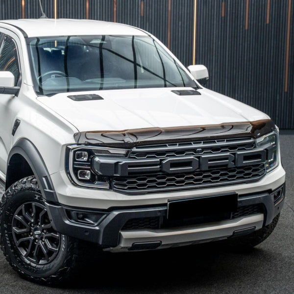 Pickup truck with "FORD" on the grille, parked in a covered area with vertical wood slat walls. White body and large tires convey a robust, contemporary design.