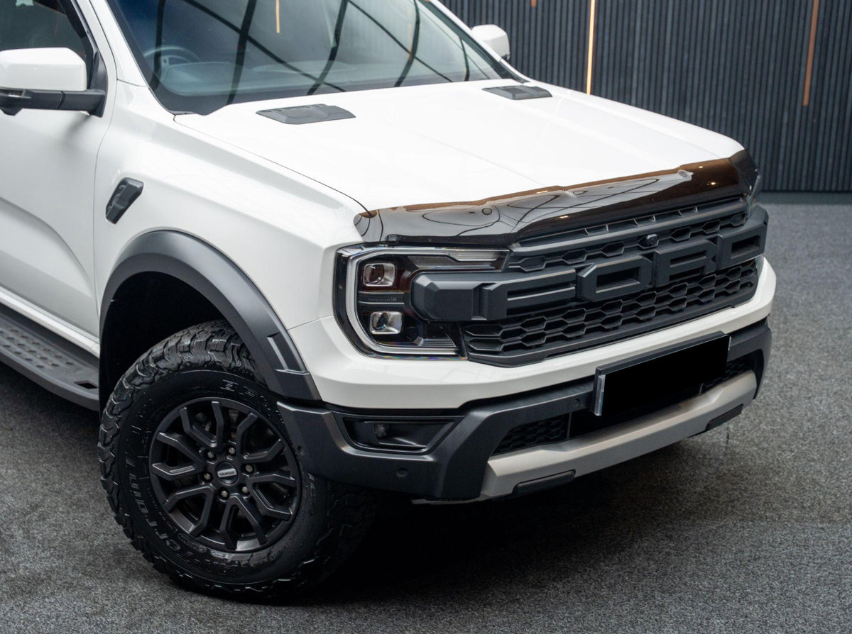 A white pickup truck with "FORD" marked on its front grille is parked on a textured surface, featuring off-road tires and a sleek black hood deflector in an indoor setting.