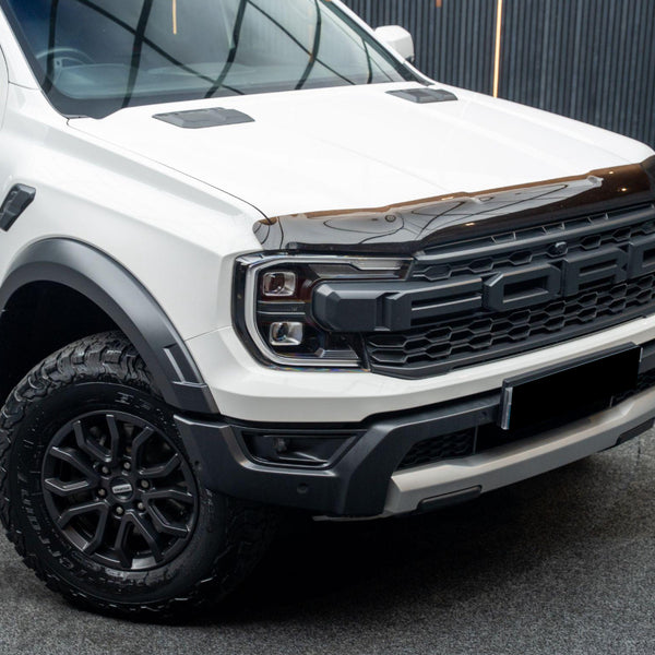 A white pickup truck with "FORD" marked on its front grille is parked on a textured surface, featuring off-road tires and a sleek black hood deflector in an indoor setting.