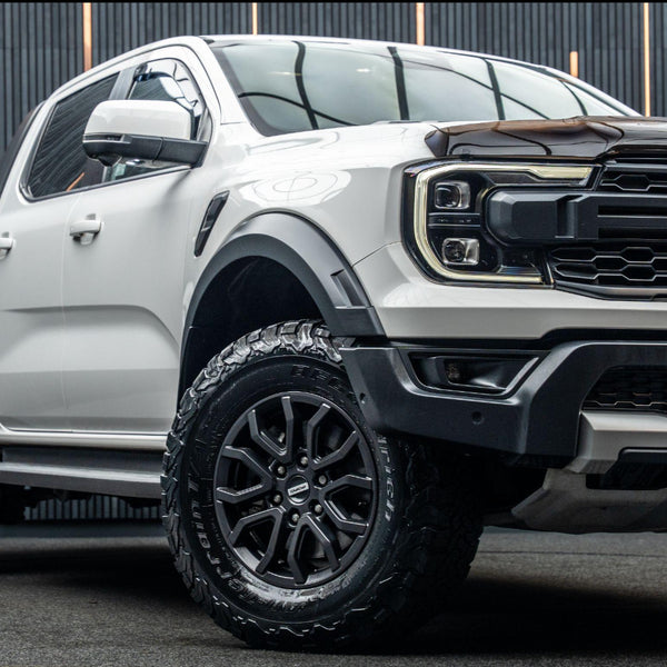 A white pickup truck with black tires is parked on a dark surface. It features prominent headlights and a rugged grill, set against a backdrop of vertical black lines.