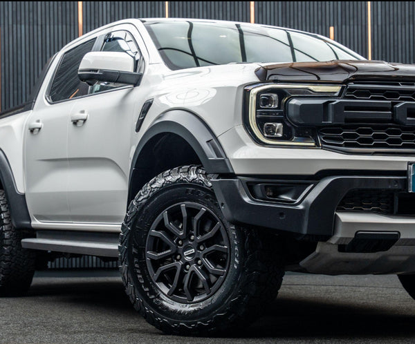 A white pickup truck with black tires is parked on a dark surface. It features prominent headlights and a rugged grill, set against a backdrop of vertical black lines.