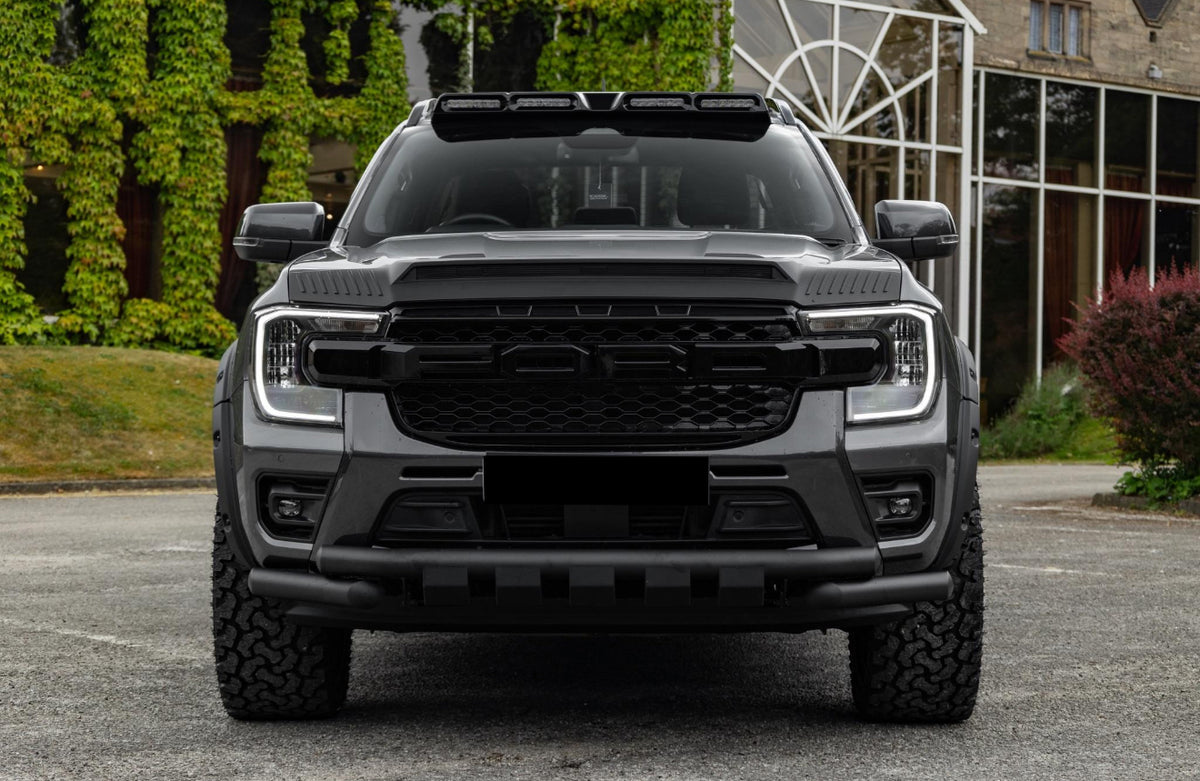A large black SUV with prominent grill and lights parked on asphalt, surrounded by greenery and a building with glass windows in the background.