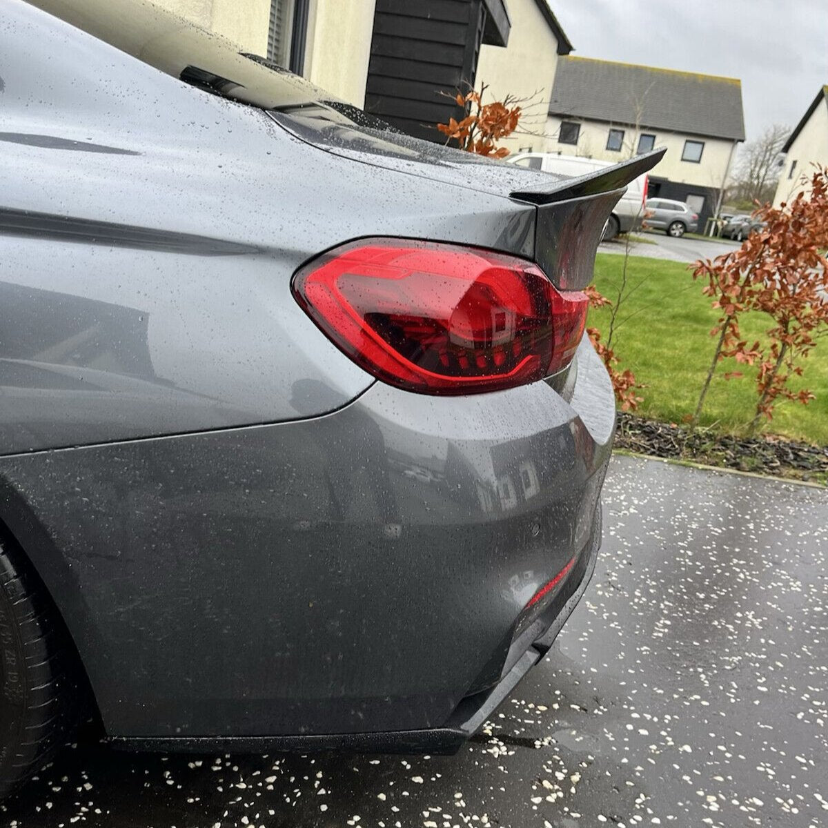 A gray car with a red taillight and a rear spoiler is parked on a speckled driveway. Nearby, brown-leaved shrubs and houses with gray roofs are visible under a cloudy sky.