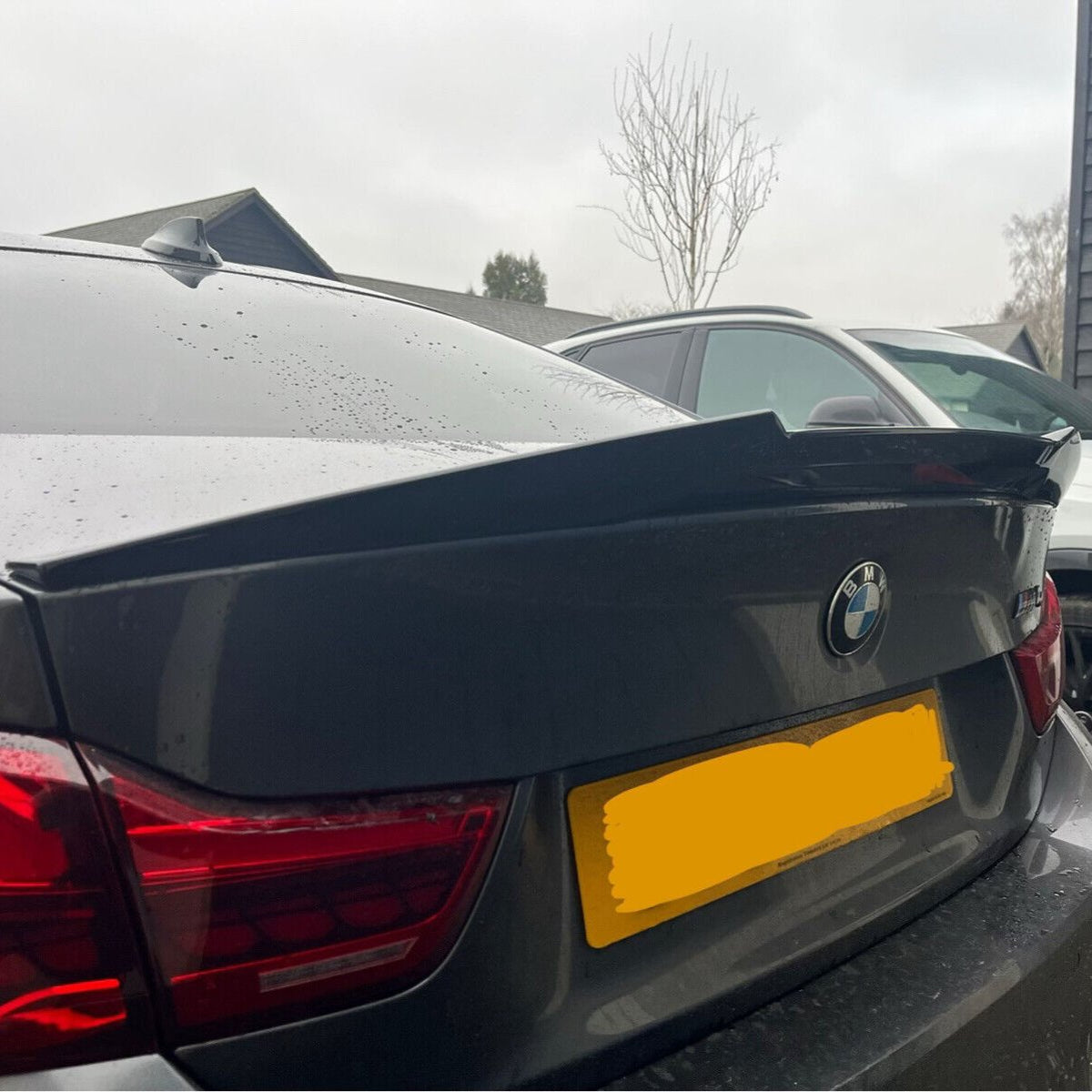 A black BMW car is parked, displaying a raised spoiler on the trunk. It's positioned in a residential driveway with trees and other vehicles nearby. The license plate is obscured.