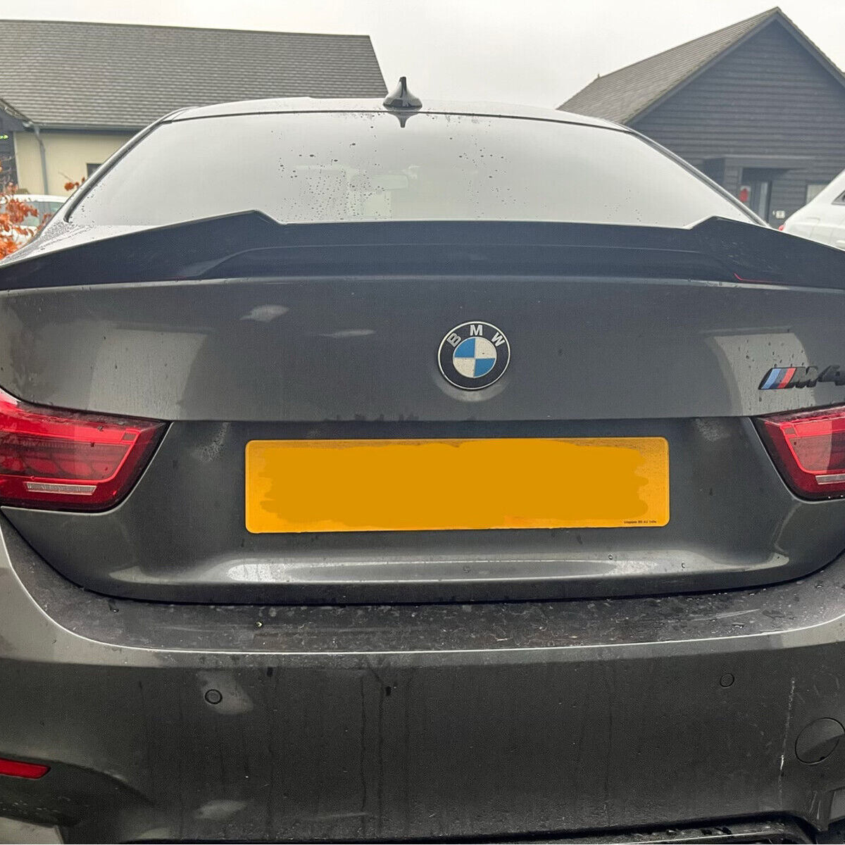 Car with "BMW" and "M4" badges parked outside, featuring a rear spoiler. The context includes residential buildings and another vehicle nearby. The license plate is yellow and partly obscured.