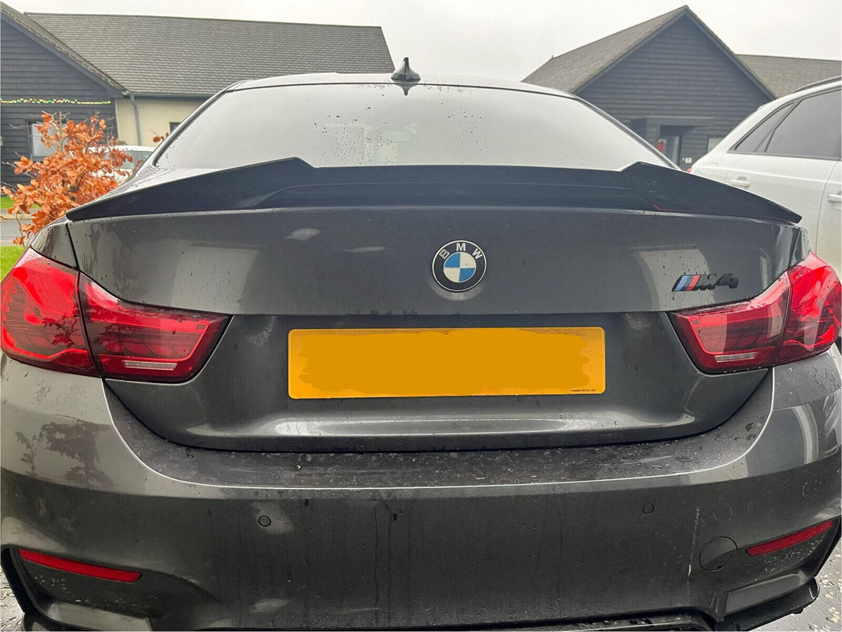 Car with "BMW" and "M4" badges parked outside, featuring a rear spoiler. The context includes residential buildings and another vehicle nearby. The license plate is yellow and partly obscured.