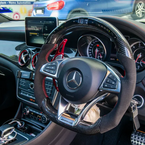 Steering wheel with multi-function controls sits inside a modern car interior, featuring a digital dashboard, infotainment screen, and climate controls. The Mercedes-AMG logo is visible on the steering wheel.