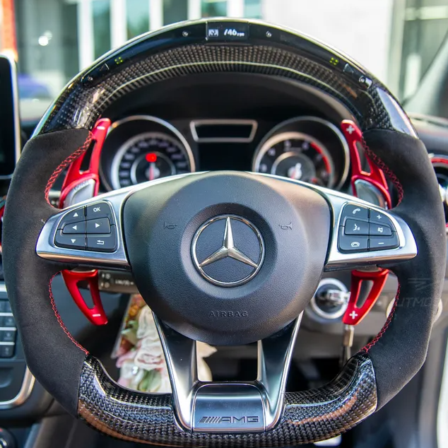 A Mercedes-AMG steering wheel with red paddle shifters, surrounded by a modern car dashboard displaying two circular gauges. The logo and buttons for audio control are visible.