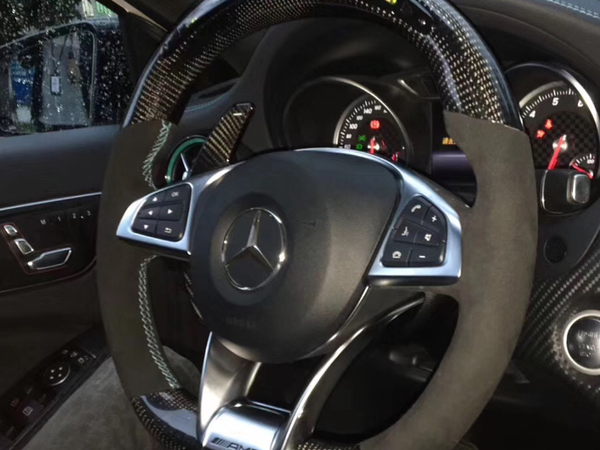 Steering wheel with carbon fiber design and buttons, displaying a Mercedes logo. Behind, illuminated speedometer gauges visible inside a car interior, with rain-covered window outside. "AMG" marked on steering wheel.