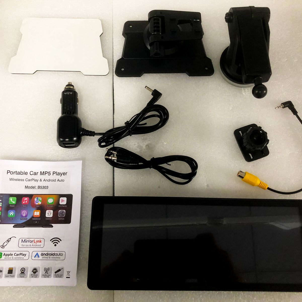 A collection of car accessories including a touchscreen, mounts, power adapter, and cables, arranged on a white foam surface. Includes manual: "Portable Car MP5 Player, Wireless CarPlay & Android Auto, Model: B5303", showing app icons.