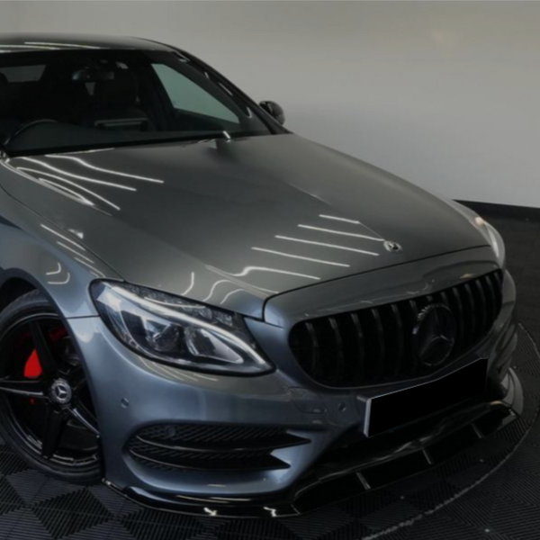 A sleek gray car parked indoors, featuring a prominent grille and headlights, with red brake calipers, on a checkered floor. The setting is a minimalist showroom.