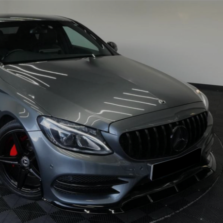 A sleek gray car parked indoors, featuring a prominent grille and headlights, with red brake calipers, on a checkered floor. The setting is a minimalist showroom.