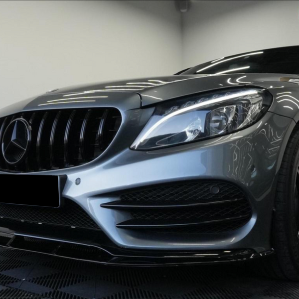 A sleek, silver Mercedes-Benz car is parked indoors on a checkered floor, showcasing its emblematic front grille, headlight design, and stylish black wheels. Bright ceiling lights reflect on the hood.