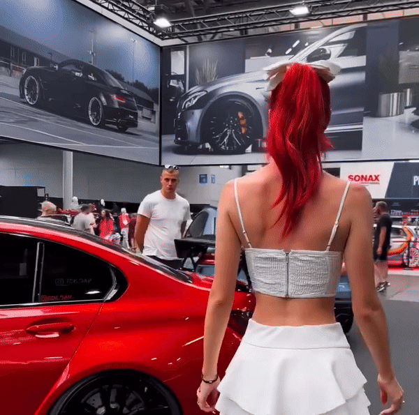 A person with red hair walks past a red car at an auto show, surrounded by large car posters and people.