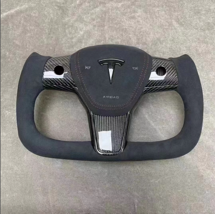Steering yoke featuring a central emblem with two horizontal buttons. Constructed from carbon fiber and wrapped in suede, situated against a plain gray background. Text reads "AIRBAG" below the emblem.