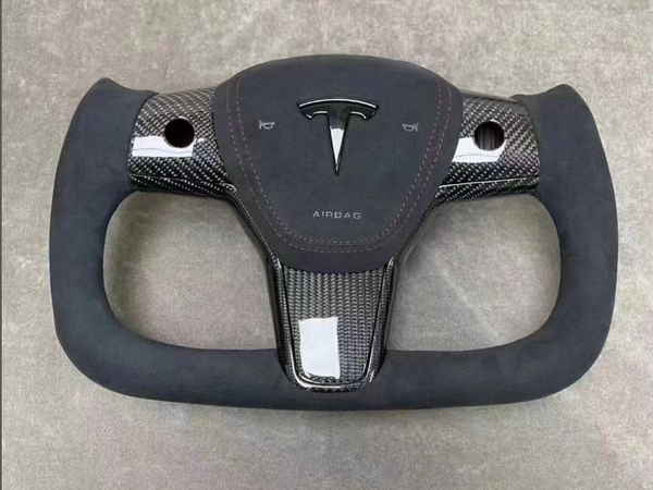 Rectangular steering wheel with a carbon-fiber design featuring the Tesla logo and "AIRBAG" text. It lies flat against a textured gray surface.