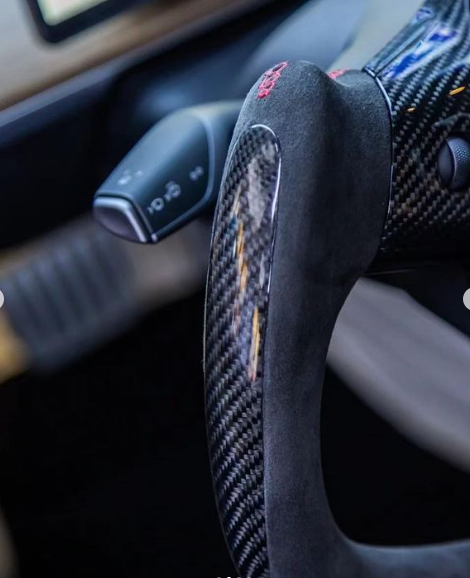 A carbon fiber steering wheel with a suede grip is in a car's interior, beside a turn signal lever. The dashboard background includes wood trim and a digital display.
