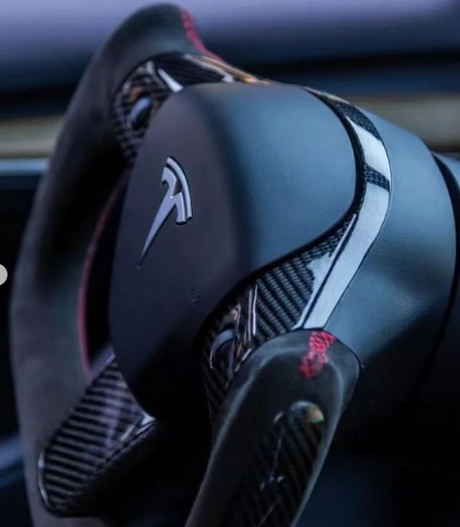 A steering yoke with a Tesla logo, accented by carbon fiber and red stitching, is situated inside a vehicle's dimly lit interior.