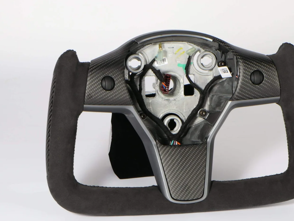 A square-shaped racing steering wheel sits against a white background. It is made of carbon fiber and suede, with visible internal components and wiring in the center area.