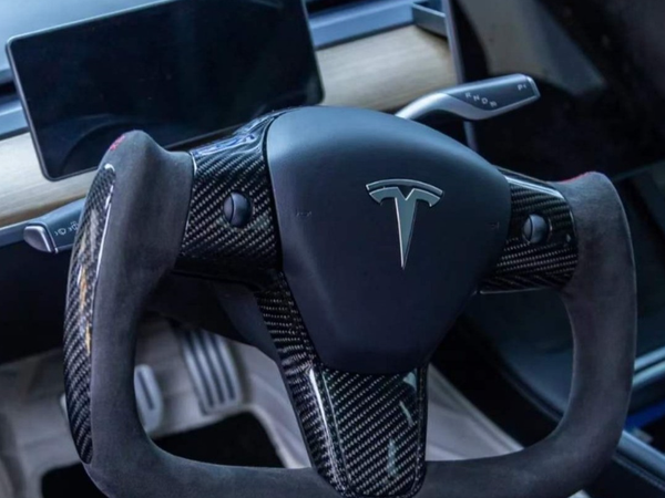 Yoke-style carbon fiber steering wheel features a Tesla logo, positioned inside a car dashboard with a touchscreen and wooden accents, surrounded by sleek, modern design elements.