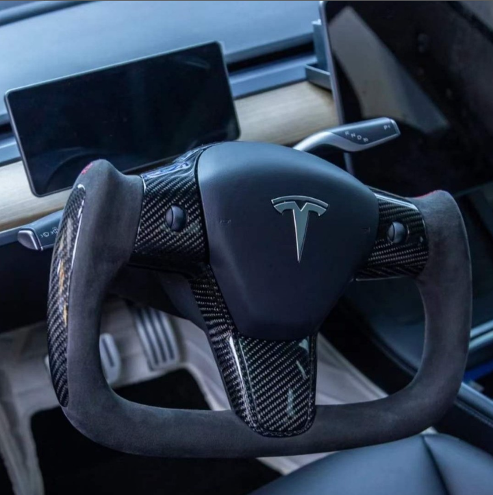 Yoke-style carbon fiber steering wheel features a Tesla logo, positioned inside a car dashboard with a touchscreen and wooden accents, surrounded by sleek, modern design elements.