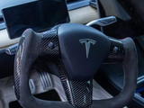 Yoke-shaped steering wheel with carbon fiber accents in a Tesla interior; dashboard display screen visible in the background, highlighting a modern, high-tech vehicle design.
