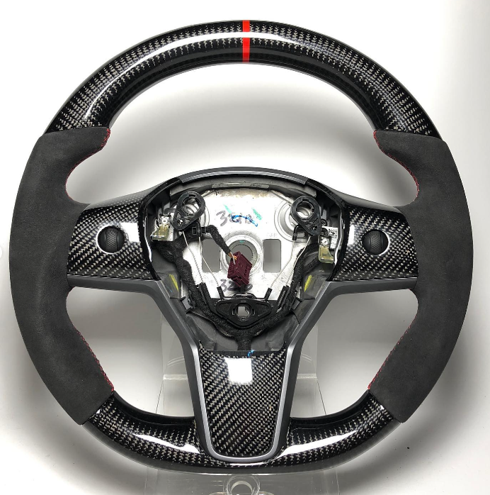 A carbon fiber steering wheel with black suede hand grips and a red top stripe is displayed on a plain white background. Center label reads "36A."