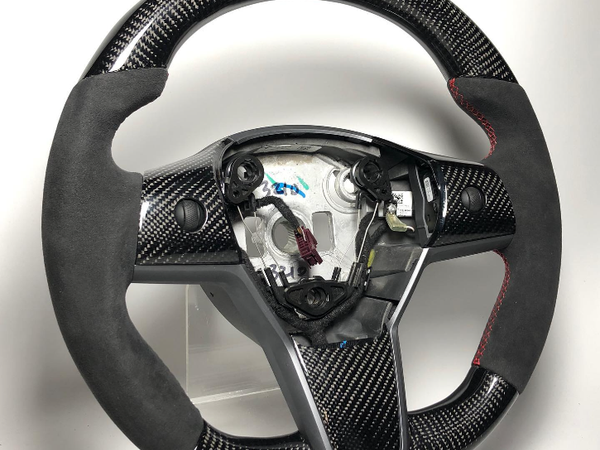 A carbon fiber steering wheel with suede grips and red stitching is displayed on a clear stand in a well-lit environment. The center is exposed, revealing internal mechanisms.