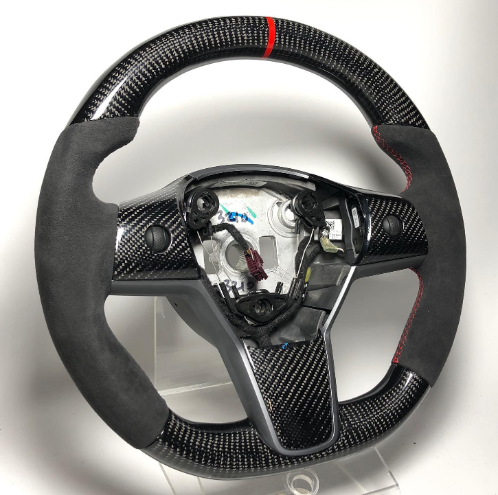 A black carbon-fiber steering wheel with suede grips and red stitching is displayed on a transparent stand, showcasing wiring and internal components in a well-lit, plain setting.